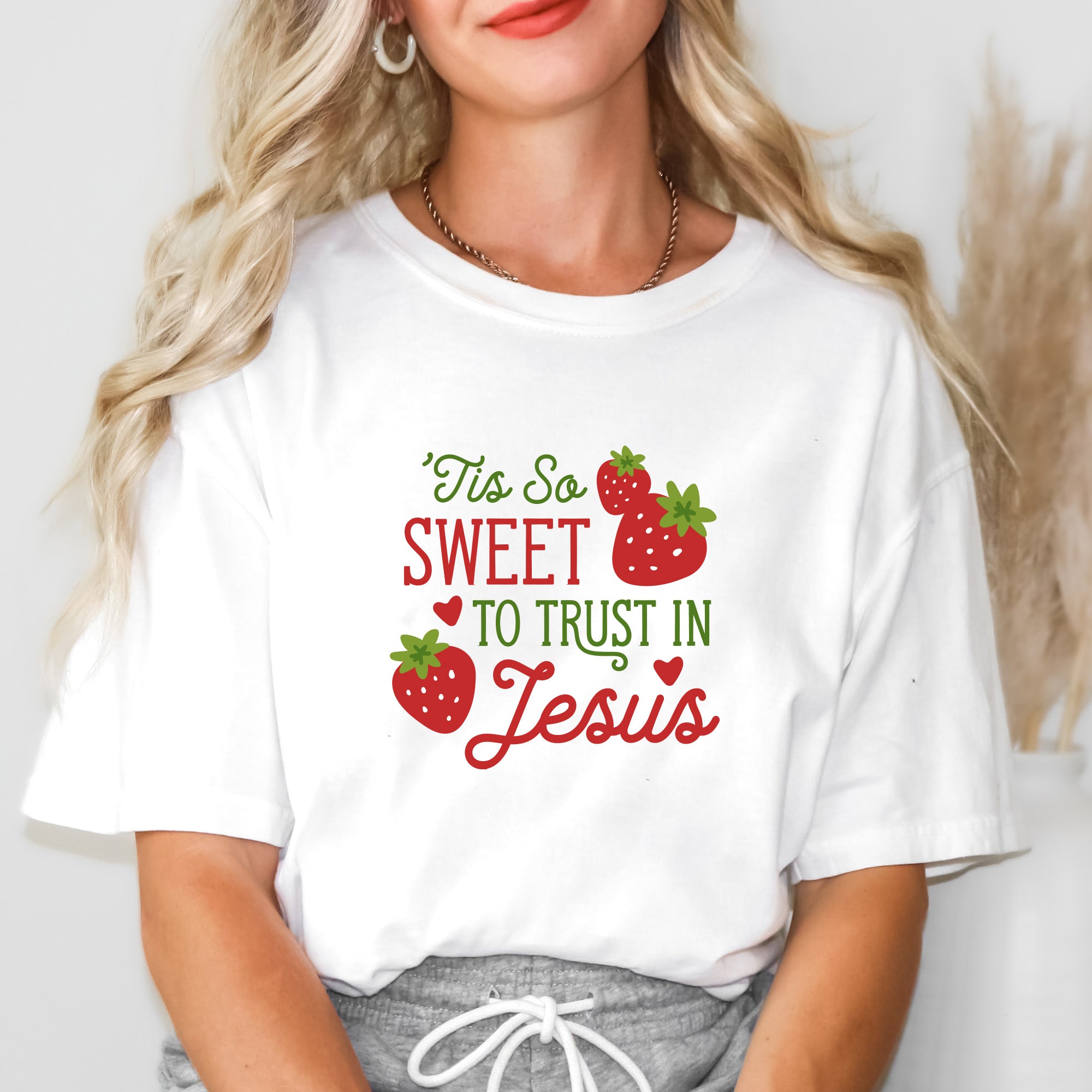 Caucasian woman with long blonde hair wearing "Tis So Sweet to Trust in Jesus" shirt in white with red and green text and red strawberries with green stems